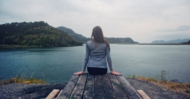 Seating - Woman Wearing Gray Long-sleeved Shirt and Black Black Bottoms Outfit Sitting on Gray Wooden Picnic Table Facing Towards Calm Body of Water at Daytime